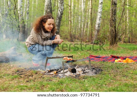 Happy woman sits near campfire with grill and barbecue in forest