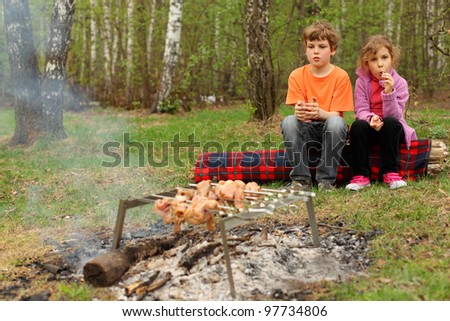 Two children sit near campfire with grill and barbecue; brother holds glass, sister sucks lollipop