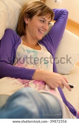 A beautiful young woman laying on her couch, smiling as she watches a funny show on television.