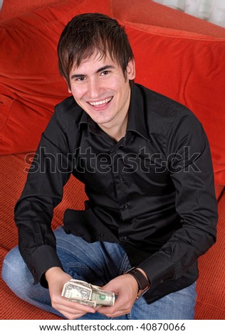 A young man seated on a bright orange sofa, wearing a black button down shirt smiling as he holds money in his hand.