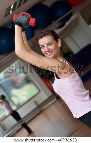 A beautiful young smiling woman standing on a wooden gym floor in front of a mirror exercising with dumbell weights raised over her head.