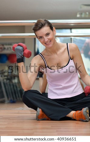 A pretty young woman exercising with dumbells, seated on a wooden gym floor in front of a glass wall. FRONT VIEW