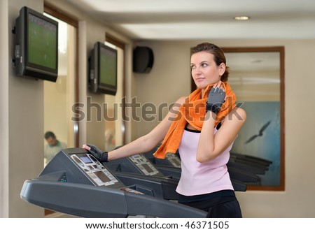 A young woman on a treadmill at the gym, wiping sweat off her neck with her orange workout towel
