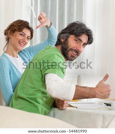 Couple are seated at their dining room table. The woman is showing a thumbs down. The man is showing a thumbs up.