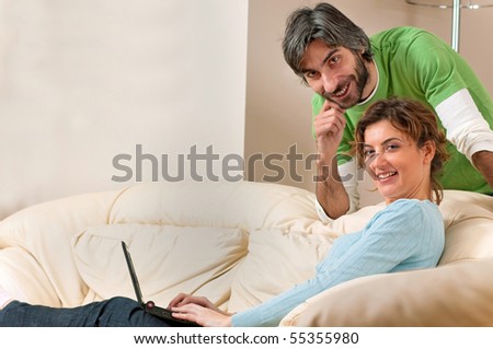 A young woman is seated on a cream couch as she types on her laptop. A man is behind the couch leaning over her. Both are posing and smiling at the camera.