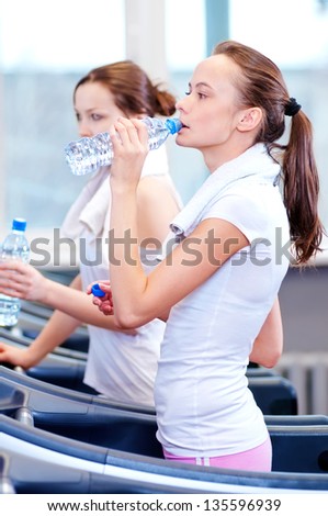 Two young women drinking water after sports. Fitness gym.