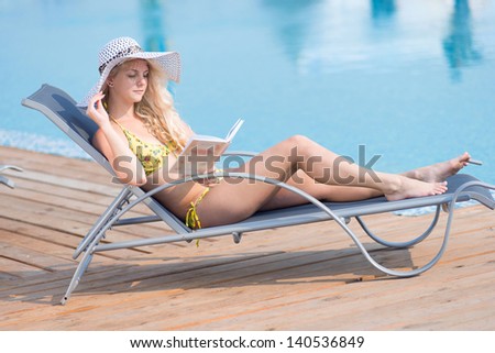 Young woman in bikini, hat and  swimsuit laying on chaise-longue and reading book by the pool in a summer vacation