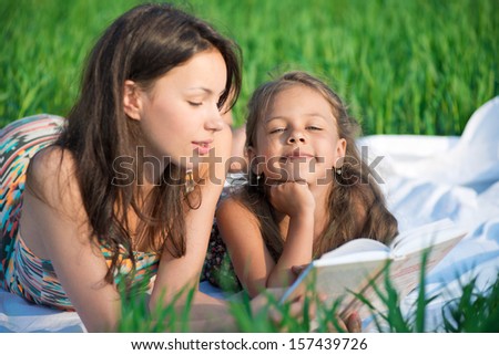 Happy girls reading book on green grass at spring or summer park picnic