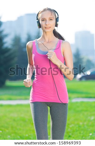 Jogging woman running in city park in sunshine on beautiful summer day and listening a music in headphones. Sport fitness model caucasian ethnicity training outdoor for marathon.