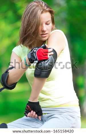Woman skating in park. Girl going rollerblading sitting on bench  putting on elbow guard protection. Sporty caucasian woman in outdoor fitness activities.