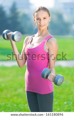 Portrait of cheerful woman in fitness wear exercising with dumbbell at green city park. Outdoors sport.
