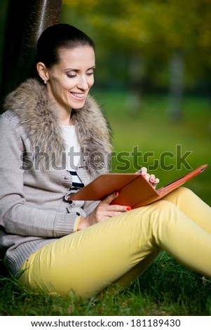 Fashion woman using a tablet computer outside in evening park. Student sitting on green grass over tree.