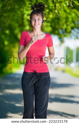 Young woman running outdoors in green park at lovely sunny summer day. Jogging with headphones and music