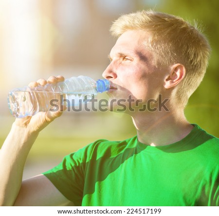 Tired man drinking water from a plastic bottle after fitness time and exercising in city street park at beautiful summer day. Sporty model caucasian ethnicity training outdoor.
