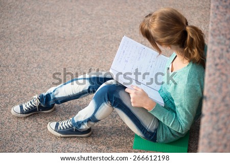 Portrait of a beautiful teenage student girl sitting on stairs in park with copy book and studing. Sunny summer day. Modern and casual lifestyle. Woman with note pad.
