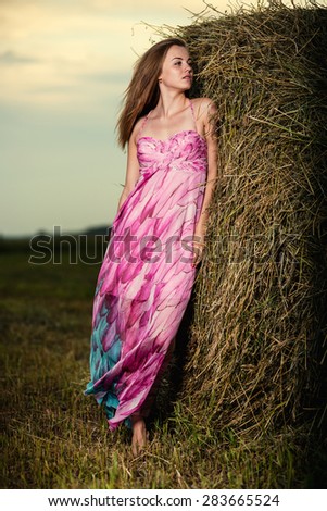 Young lady standing in evening field. Beautiful woman posing at the old rural farm location. Outdoor summer portrait of pretty fashion style woman in colored dress over haystack. Beautiful slim girl