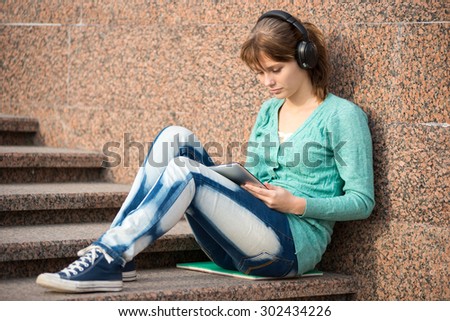 Preparing to exams outdoors. Headphones music. Beautiful young female student writing or reading something from note pad. Woman sitting on stairs in city park.