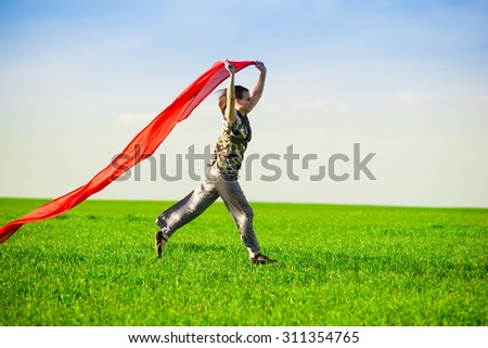 Beautiful young woman jumping on a green meadow with a colored red tissue. Happy female in wheat field with fabric. Summer picnic and freedom concept.