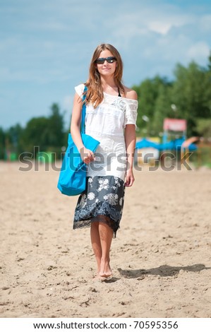 Beautiful young woman in dress and sunglasses walking on sand beach with bag on sunny summer day