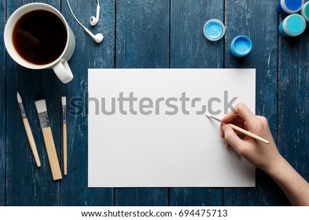 Top view of woman hand with painting brush, watercolor paints and blank sheet of paper, black coffee cup, headphones. Blue wooden background.