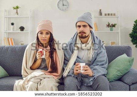Dissatisfied young couple having problem with central heating, sitting on sofa at home, freezing, drinking hot tea trying to warm up. Sick man and woman wrapped in blankets suffering from cold or flu