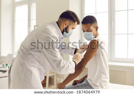 Doctor in medical face mask giving injection to patient during vaccination campaign at the clinic. Young African-American man getting Covid-19, AIDS or flu antivirus vaccine shot at the hospital