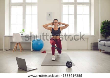 Smiling athletic young woman training legs at home watching active sports workout video lesson. Fit female athlete in activewear doing forward lunges exercise with elastic resistance rubber glute band