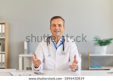 Male family doctor sits in front of the camera and gives an online consultation to his patient while sitting in the office. Friendly doctor in a medical gown explains the patient's diagnosis. Foto stock © 