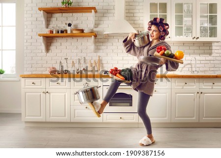 Tired housewife cooking food and carrying lots of stuff. Frail slim young woman making meal at home, holding multiple heavy cooking pots and kitchen saucepans, balancing cutting boards with vegetables