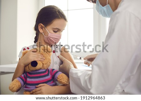Vaccine against coronavirus for kids during outbreak concept. Little girl patient in medical protective mask with toy sitting getting vaccination injection with syringe against covid-19 from doctor