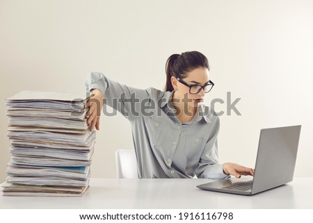 Young accountant working on laptop computer sitting at desk with pile of papers. Paperwork vs electronic documents. Storing files in digital database. Having quick convenient access to storage system