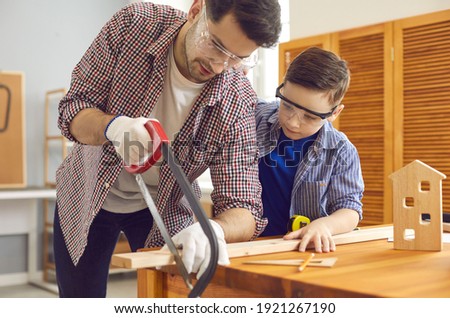 DIY. Dad teaches his little interested son to build a wooden toy house in the workshop. Concentrated father saws a wooden board while his son watches. Man teaching his son to work with wood and tools.