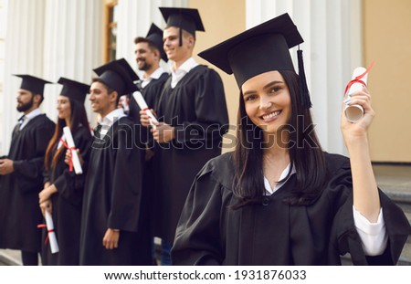 Young smiling girl university graduate standing holding diploma in raised hand over group of mates and university building at background. Graduation from university, education, diploma concept