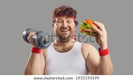 Funny smiling man with dumbbell and delicious burger looking at camera. Happy fat guy holding free weights and eating big yummy hamburger. Sport, food, failed diet, workout exercise, cheat day concept
