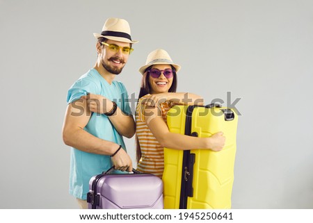 Young people ready for safe summer holiday journey. Studio portrait of happy couple holding travel cases, smiling and showing arms after receiving COVID-19 vaccine. Coronavirus vaccination concept
