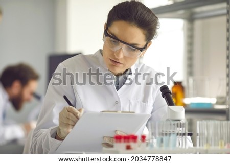 Portrait of serious scientist in white lab coat collecting data in medical research laboratory. Focused med student holding clipboard and taking notes summarising information for scientific report Imagine de stoc © 