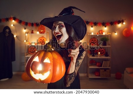 Portrait of woman in wicked witch costume. Beautiful lady in black pointed hat with happy excited face expression standing in dark room, holding Halloween jack-o-lantern pumpkin, shaking finger at you Foto stock © 