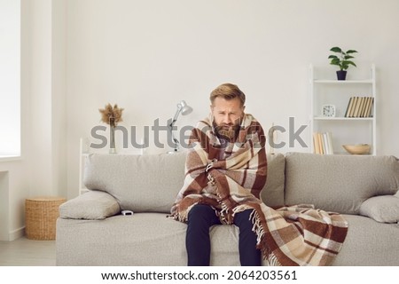 It's cold at home in wintertime. Man freezing in his house in winter because of broken thermostat. Sad bearded young guy wrapped in woolen plaid shivering while sitting on sofa in living room interior Foto stock © 