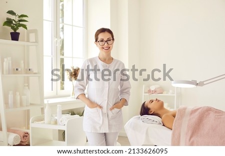 Portrait of friendly female woman beautician, aesthetic nurse or masseuse at her workplace. Smiling female beauty salon worker standing next to her client lying on couch in bright office. Foto d'archivio © 