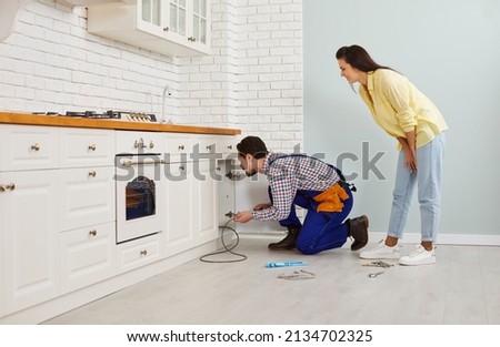 Plumber crouching on the floor in a modern white kitchen and using a drain cable to clean a clogged sink pipe. Young girl calls the plumber to fix the problem of a clogged drain under her kitchen sink Foto stock © 