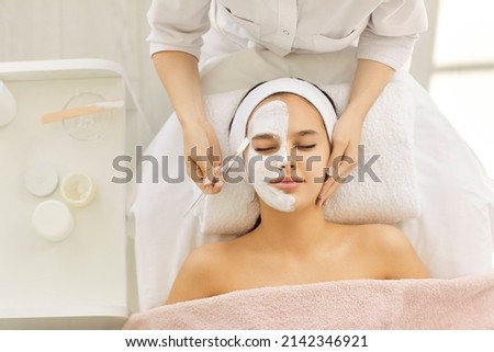 Satisfied young Caucasian beautiful woman uses services of professional beautician in beauty salon or spa. Unrecognizable beautician applied white mask to half of client's face with brush. Top view. Foto d'archivio © 