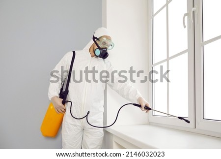 Pest control exterminator working inside house. Young guy wearing protective white suit and mask holding sprayer bottle and spraying insecticide over window sill in modern living room interior at home Zdjęcia stock © 