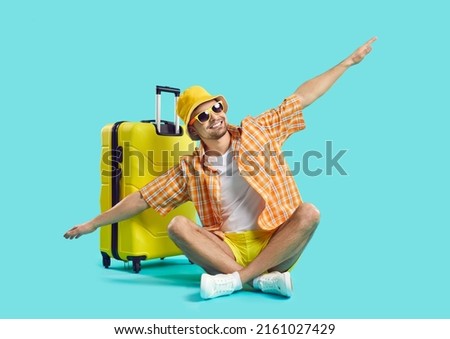 Happy man who goes on summer vacation by air poses with suitcase on light blue background. Guy in summer clothes sits with outstretched arms imitating flight of airplane. Air flight journey concept. Сток-фото © 