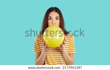 Woman blowing up birthday party balloon. Young brunette girl in striped orange white tee shirt isolated on turquoise background, inflating yellow balloon and looking at you with funny face expression Сток-фото © 