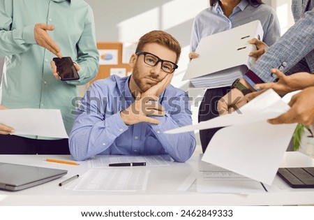 Stress, burnout and tired man sitting at the desk in office frustrated or overwhelmed by coworkers at workplace. Young businessman looking sad at camera. Overworked, multitasking concept.