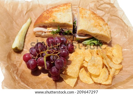 A picnic lunch with a Turkey and Cheese Sandwich on Cheese Bread, Chips, Red Grapes and a Dill Pickle Slice wrapped in deli paper, isolated on white. Deli Sandwiches are a favorite for picnics