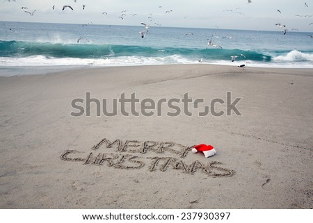 The words Merry Christmas written in the sand with a Red Christmas Santa Claus hat next to the word Merry with the Ocean and Seagulls in the background. Christmas is the time many go to the beach