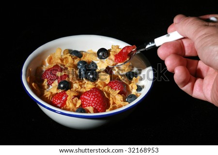 low key studio shot of hand holding a spoon full of cereal in the foreground and a bowl of cereal with strawberries and blueberries in the background isolated on black
