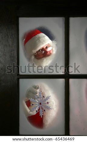 Santa Claus looks out through the snow and fog on his workshop window in the north pole to see outside check the weather while he holds up a large snow flake  on December 24th, Christmas Eve