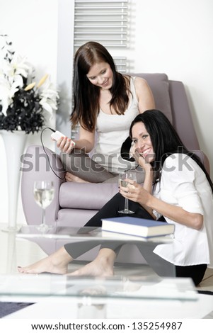 Two beautiful young woman listening to music on headphones at home on a chair.